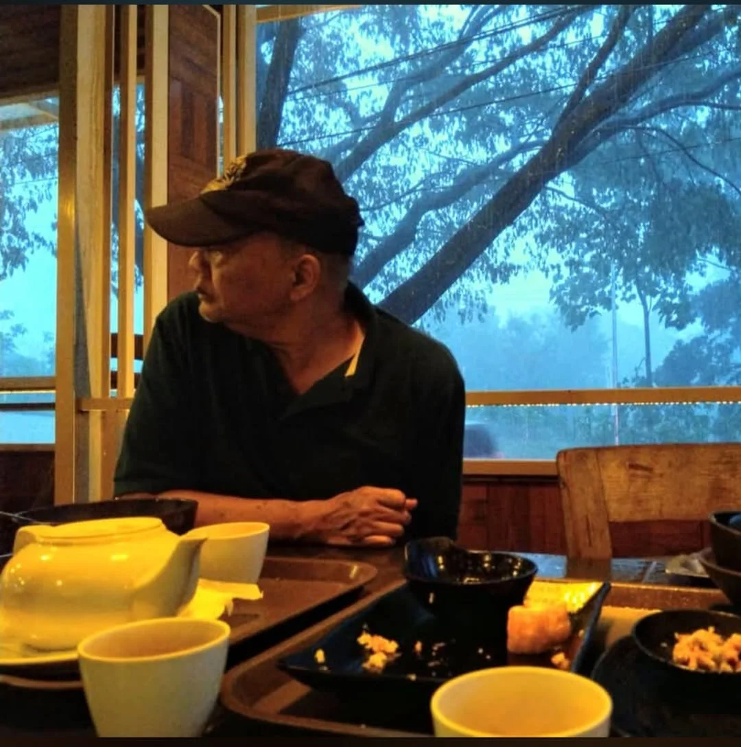 Seorang pria mengenakan topi hitam duduk di meja makan dengan pemandangan pohon-pohon besar melalui jendela kaca di belakangnya.