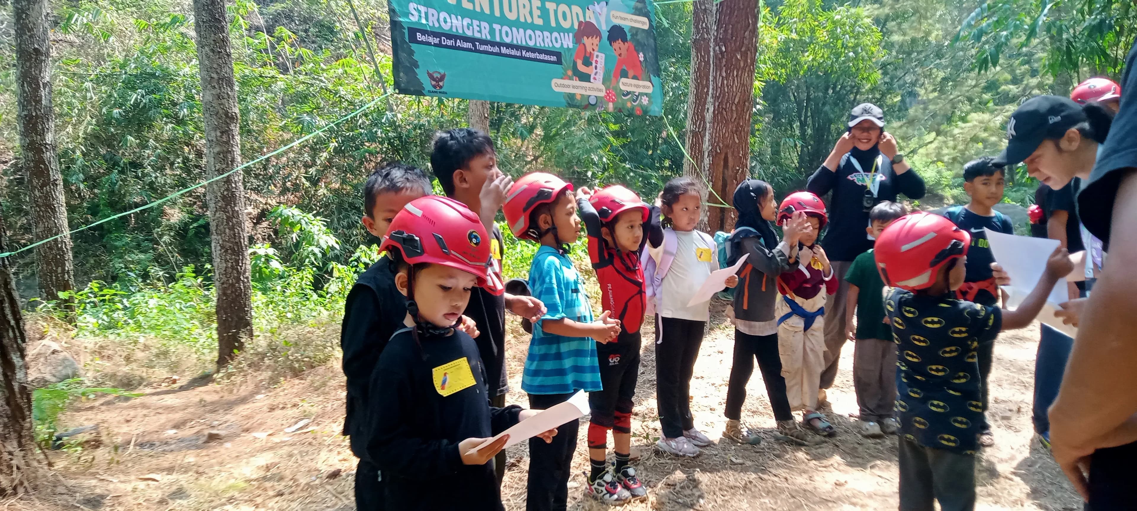 Sekelompok anak-anak mengenakan helm merah berkumpul di jalur hutan selama kegiatan petualangan outdoor yang dipandu oleh instruktur.